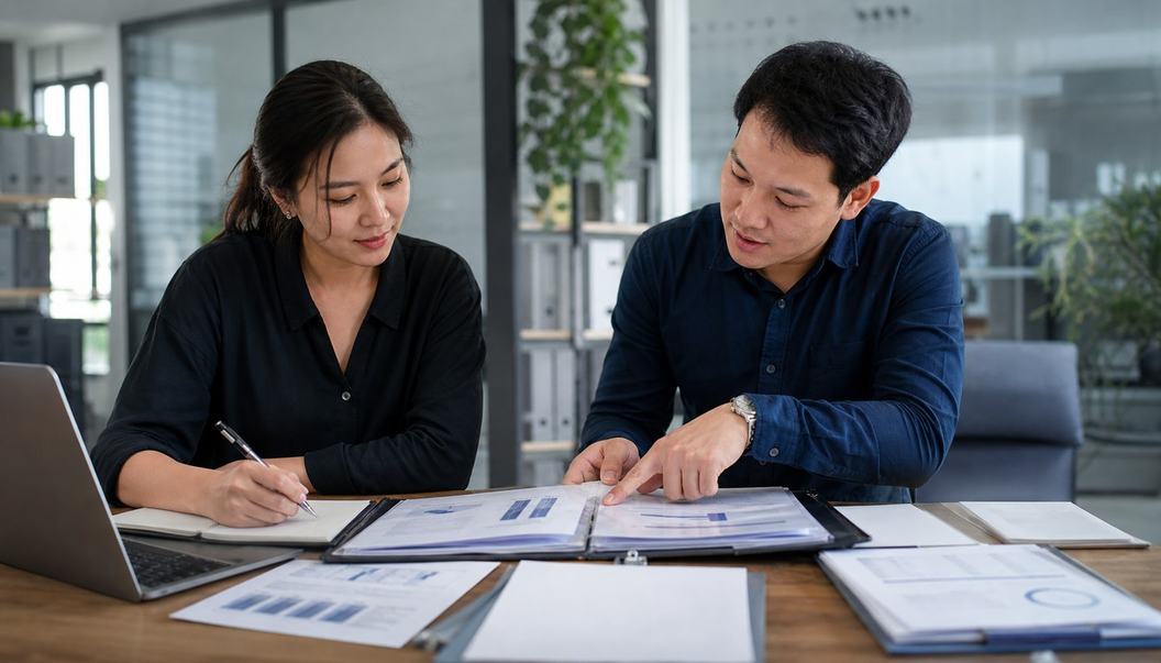 A man points to a page in an open binder while a woman takes notes beside a laptop and loose business papers.
